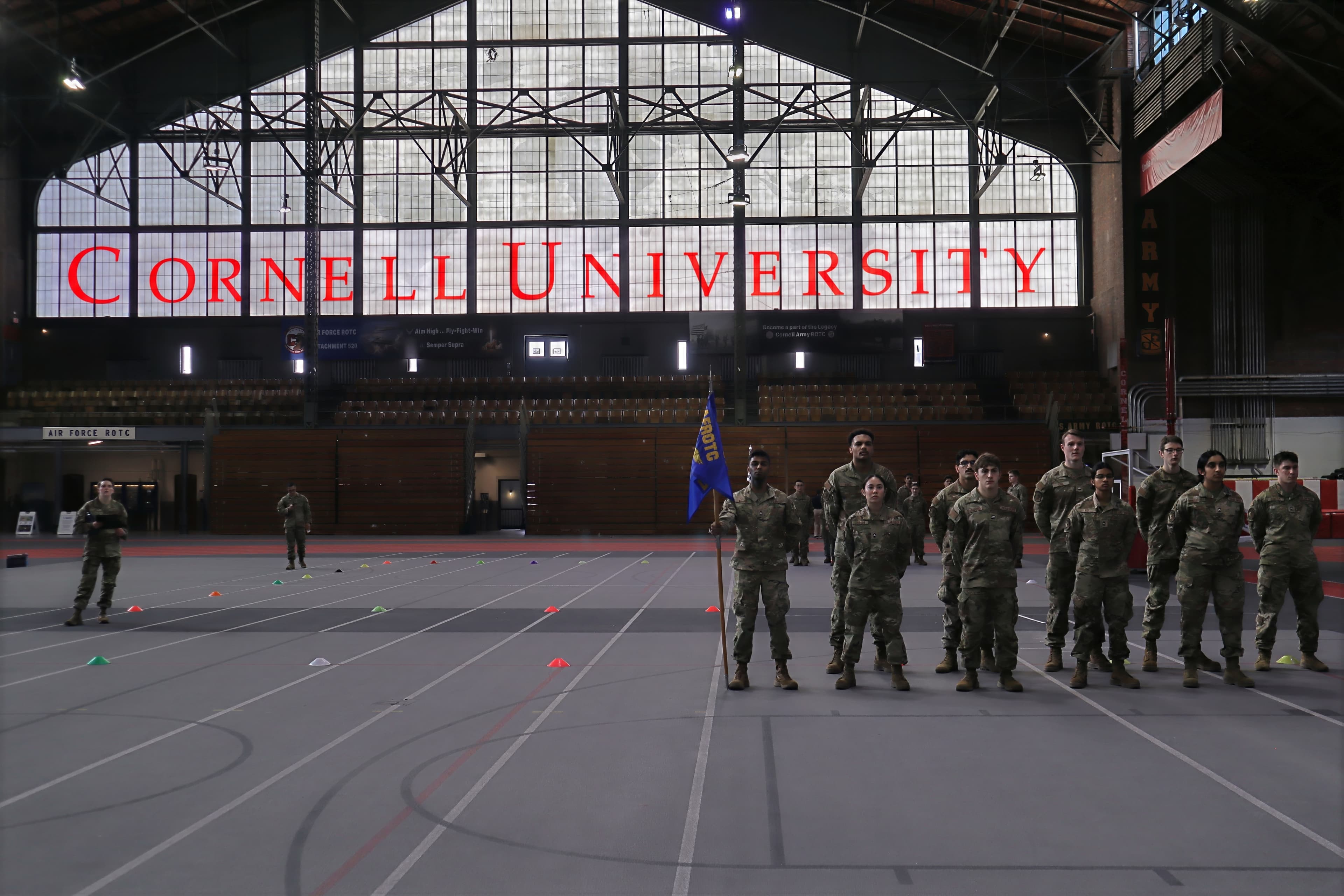 Detachment 520 cadets in formation with their guidon on the Barton Hall floor beneath tall windows lettered Cornell University.