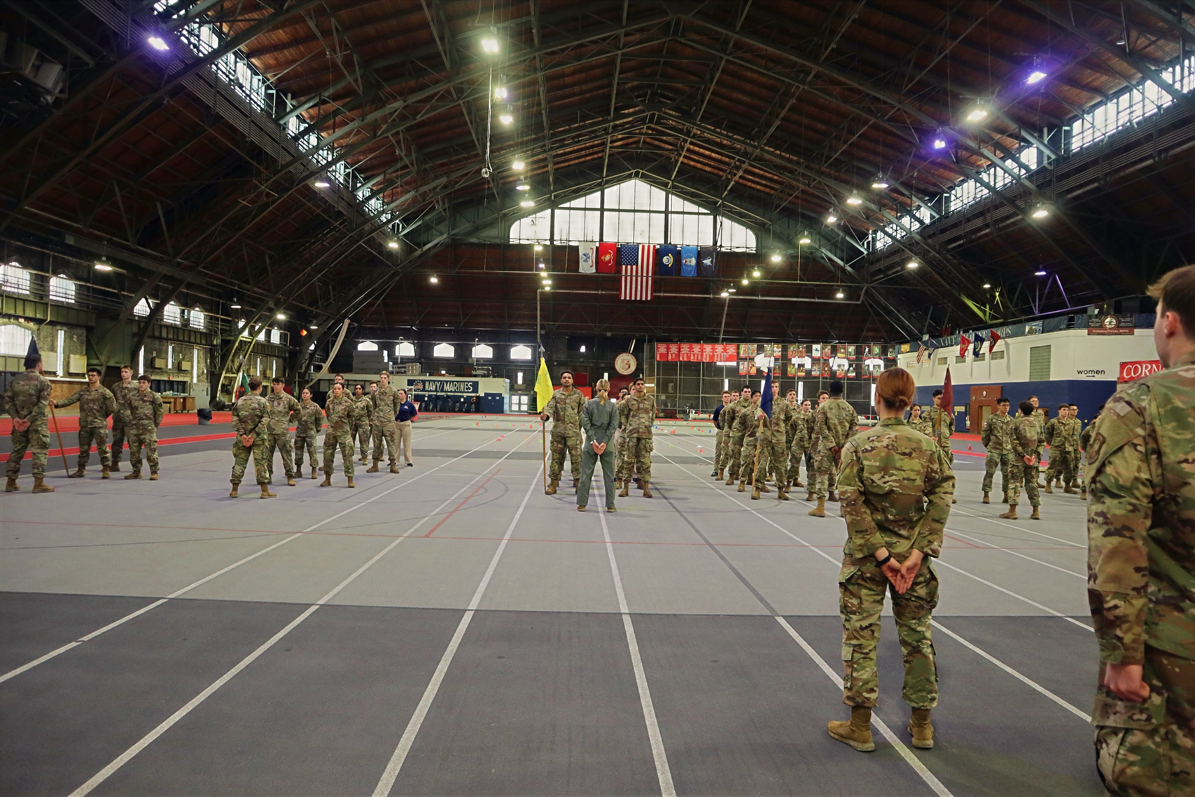 The cadet wing stands in formation across the Barton Hall floor, faced by its commander, with the national and service flags hanging above.
