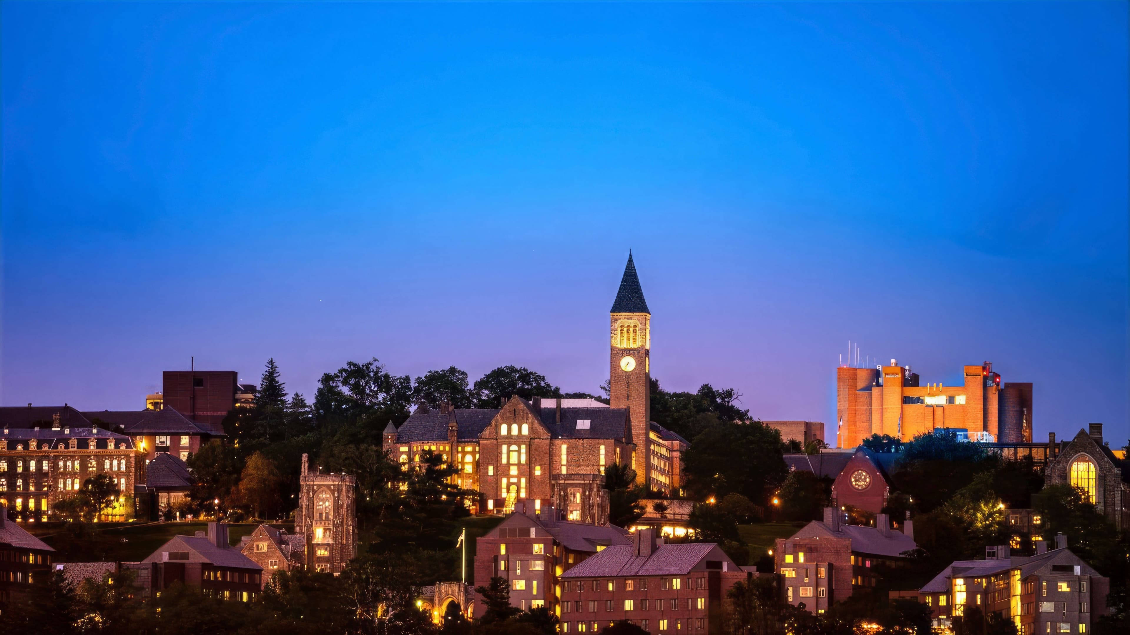 Cornell's campus skyline at dusk, with McGraw Tower and surrounding buildings lit against a deep blue sky.